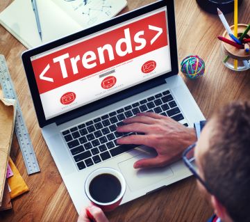 A view looking over the shoulder of a person working at a laptop and holding a cup of coffee. The screen on the laptop has the word TRENDS in large white letters on a red background.